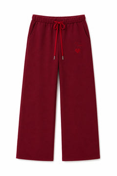 A pair of wide-leg sweatpants in deep burgundy red, featuring an elastic waistband with a matching red drawstring and a small embroidered red heart on the left thigh, shown against a plain white background.