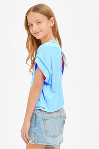 A girl wearing a neon coral sleeveless boxy crop top with a crew neckline and visible contrast overlock stitching along the armholes, side seams, and hem, paired with light-wash denim shorts.