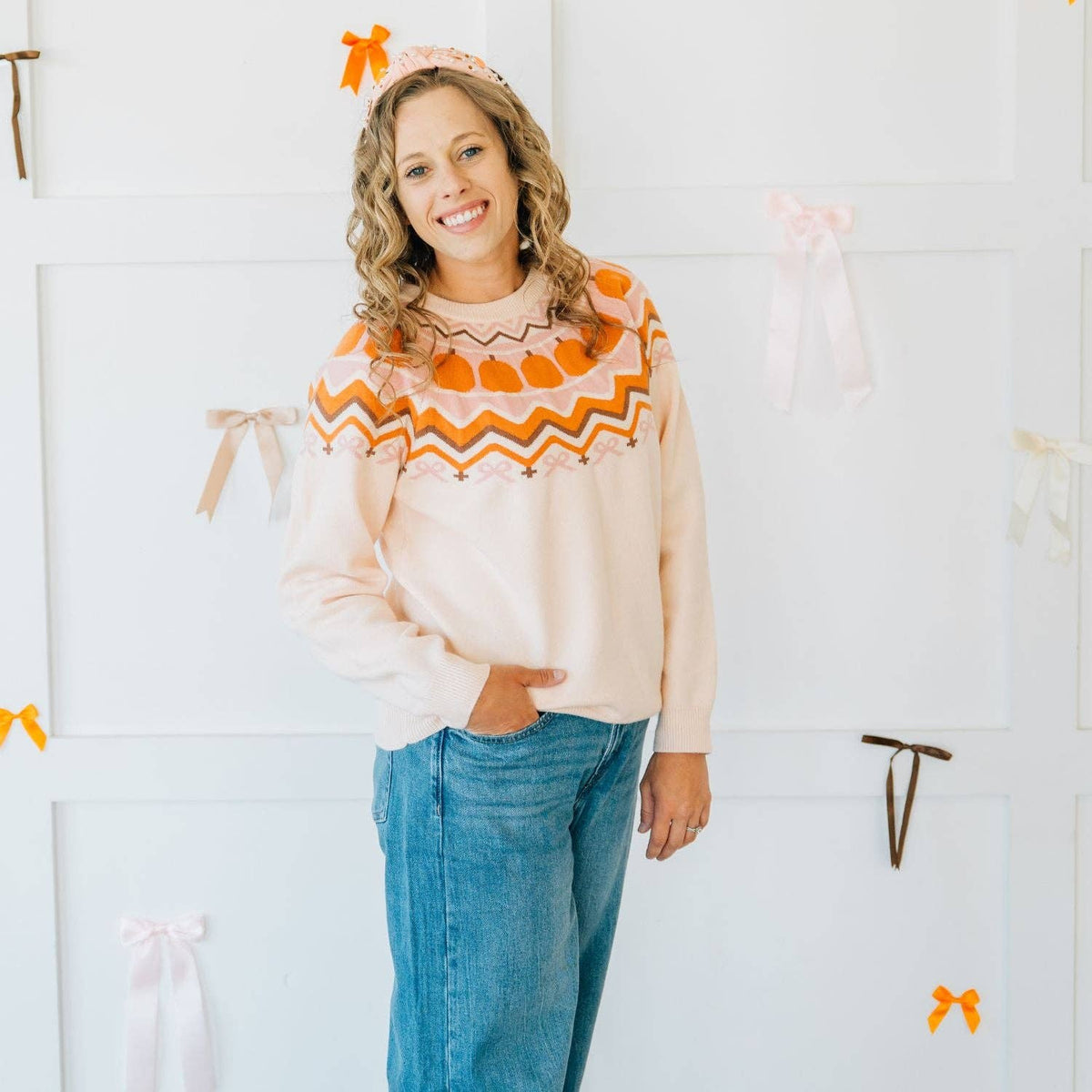 Woman wearing a cream sweater with orange patterns and blue jeans, standing against a white wall.
