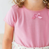 A close-up of a child wearing a light pink short-sleeve top with delicate pink scalloped trim around the neckline and sleeve cuffs, plus a small pink bow at the center chest.