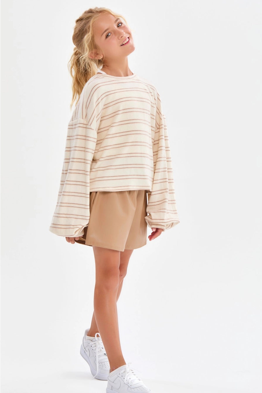 A girl wearing a cream and taupe fine horizontal stripe brushed knit pullover top with a crew neckline, long sleeves with slightly flared cuffs, and a relaxed fit.