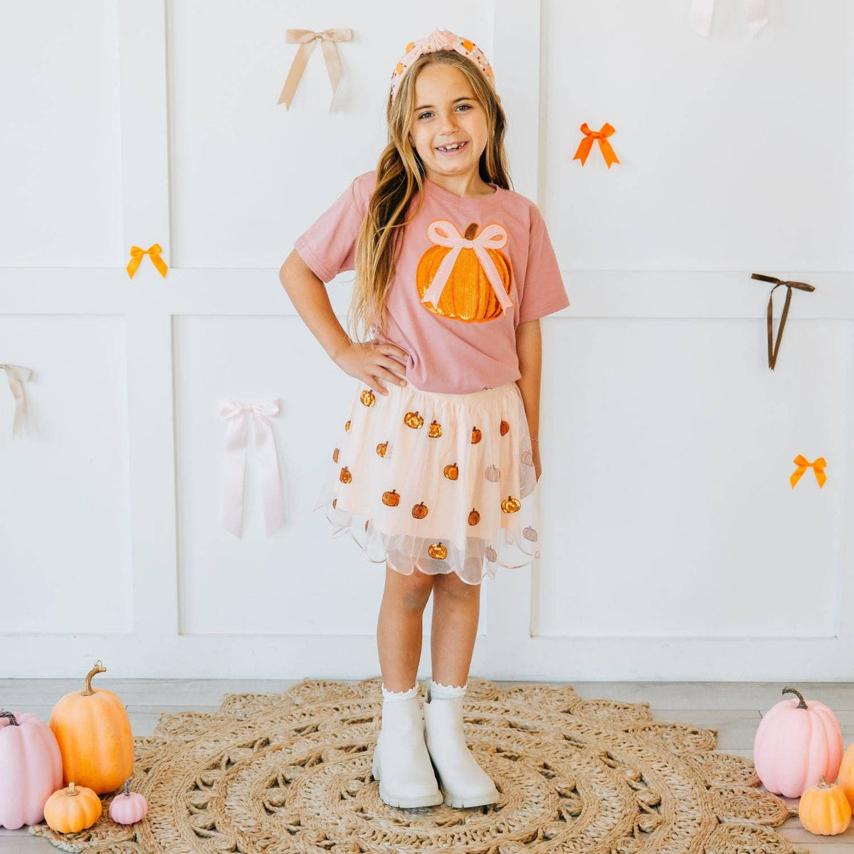 Young girl in a pink shirt with a pumpkin design and a matching skirt standing in a room with decorative bows and pumpkins.
