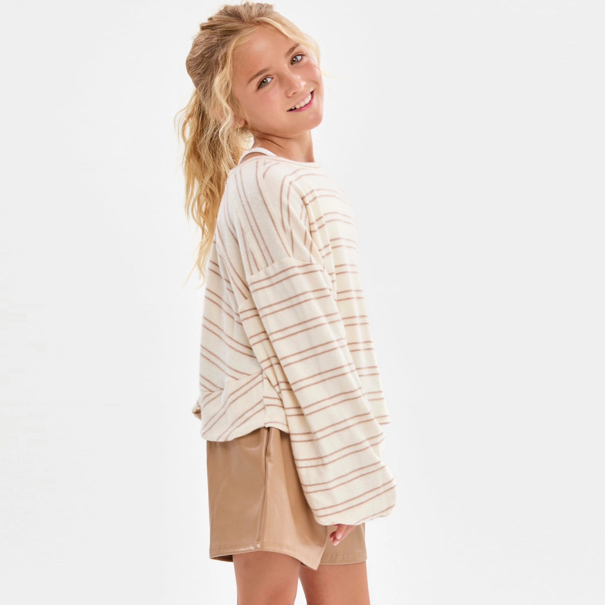 A girl wearing a cream and taupe fine horizontal stripe brushed knit pullover top with a crew neckline, long sleeves with slightly flared cuffs, and a relaxed fit.