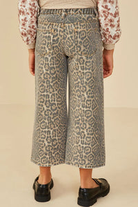 Back view of a person wearing wide-leg cropped denim jeans with an allover leopard print pattern in beige, gray, and brown tones, paired with a floral long-sleeve top and black loafers, against a plain beige studio background.