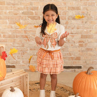 A young girl smiling and playing with falling autumn leaves, wearing a white t-shirt with an orange gingham bow design and a matching orange gingham pleated skirt. She is standing in a fall-themed setting with pumpkins and a brick wall background.
