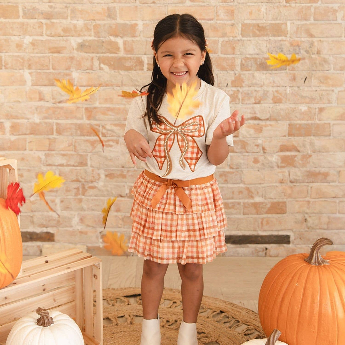 A young girl smiling and playing with falling autumn leaves, wearing a white t-shirt with an orange gingham bow design and a matching orange gingham pleated skirt. She is standing in a fall-themed setting with pumpkins and a brick wall background.