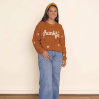 A woman wearing a rust-orange ribbed knit sweater with 'thankful' written across the chest in white script, paired with light-wash blue jeans and a matching orange headband, standing against a plain cream wall.