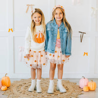 Two young girls in matching outfits with pumpkin designs standing on a rug with pumpkins around.