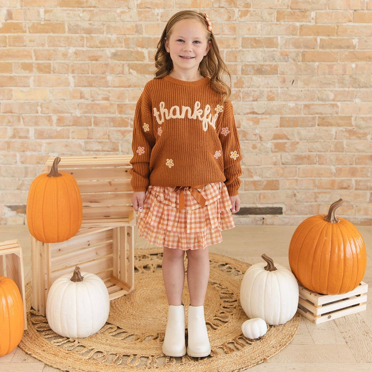 A young girl stands in a fall-themed room wearing a rust-orange ribbed knit sweater with 'thankful' embroidered on the chest, a peach and white gingham skirt, and white ankle boots. She is posed on a woven rug with pumpkins and wooden crates against a brick wall.