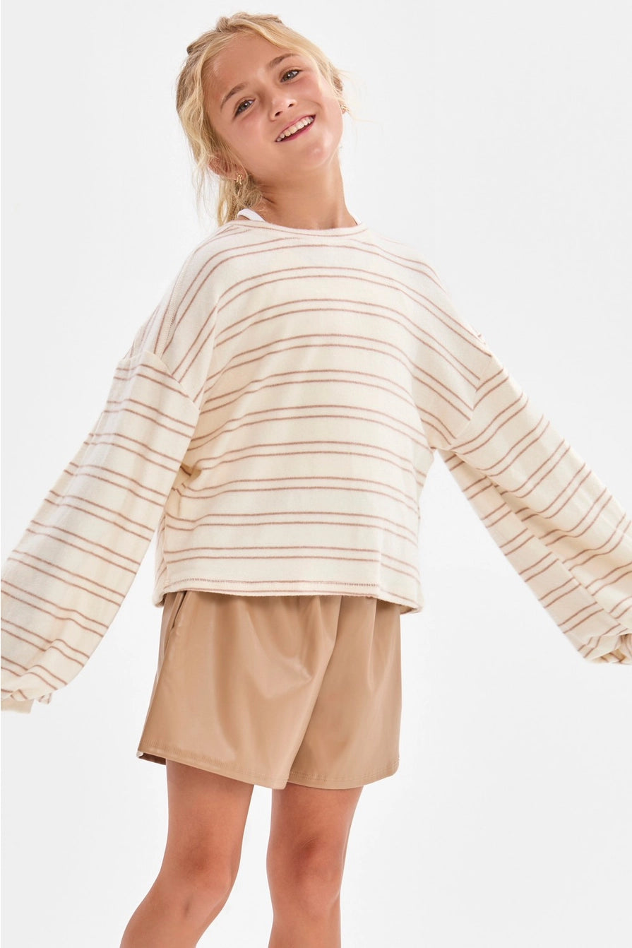 A girl wearing a cream and taupe fine horizontal stripe brushed knit pullover top with a crew neckline, long sleeves with slightly flared cuffs, and a relaxed fit.